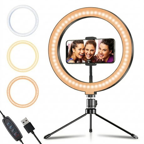 A 10-inch LED ring light mounted on a black tabletop tripod, shown at a slight angle on a white background. The ring light is illuminated with a warm glow, featuring a smartphone held horizontally in its center. To the left, three smaller circular icons demonstrate the different light color modes, while a USB power cable with an integrated remote control extends from the bottom.
