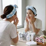 Woman applying skincare wearing coral fleece bow headband in bathroom mirror