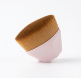 A pink flat-top foundation brush is shown on a white background, demonstrating the dense brown synthetic bristles. The brush is slightly angled, showcasing the unique shape of the bristles.op