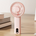 Pink handheld mini fan with display placed on a desk with books in the background. Features a digital display showing battery level.