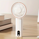 White handheld mini fan with display standing on a desk near books. The screen shows battery percentage for easy monitoring.