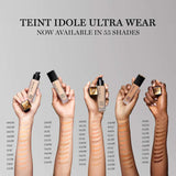 Hands holding Lancome Teint Idole Ultra Wear Foundation bottles with multiple shade swatches on arms. Displays a wide range of tones available.