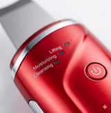 A high-resolution, macro close-up of the red ultrasonic facial cleansing device's control panel. The shot emphasizes the metallic texture and clear mode indicators for cleansing, moisturizing, and lifting.